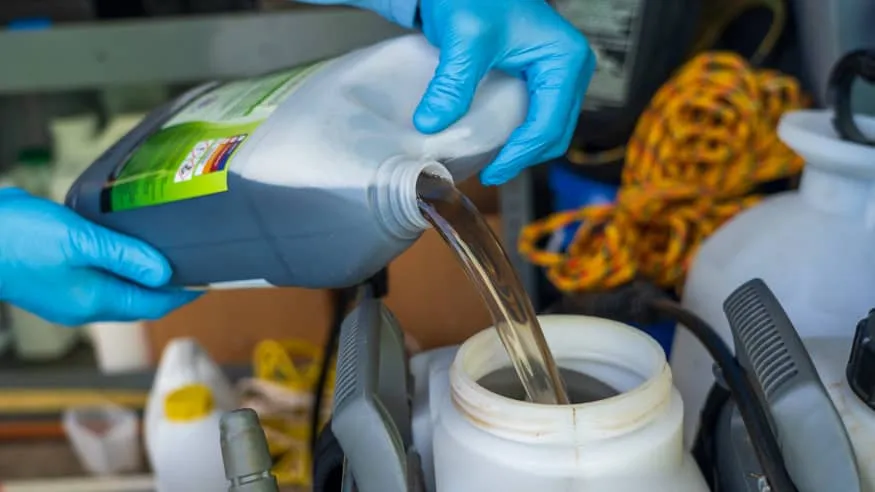 Gloved hands pouring dark brown PondZilla Pro from its jug into the open tank of a backpack sprayer, with spray equipment and hose coiled behind.